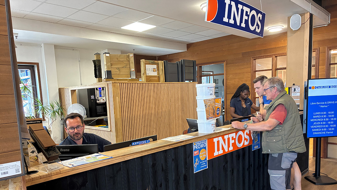 Comptoir d'information, mais cette fois avec une vue plus large de l'espace environnant. On peut voir deux employés derrière le comptoir, l'un d'eux aidant un client. Des produits en bois sont empilés derrière le comptoir, et des affiches promotionnelles 